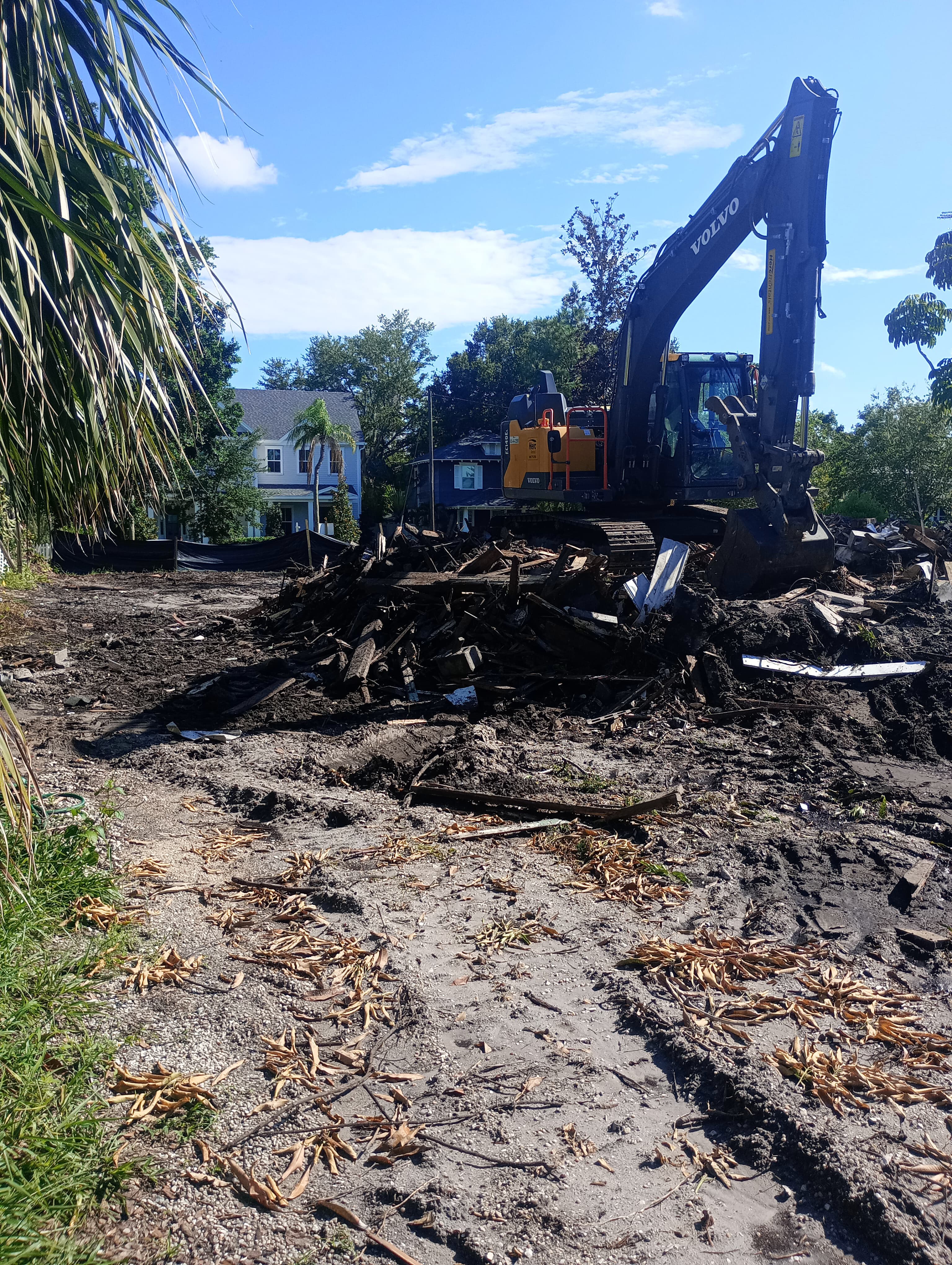 Volvo excavator clearing a pile of wooden debris at a sunny residential demolition site.