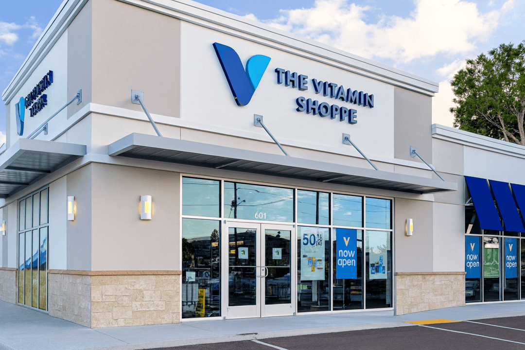 Modern Vitamin Shoppe storefront with blue logo, glass entrance, and "Now Open" signs in windows.