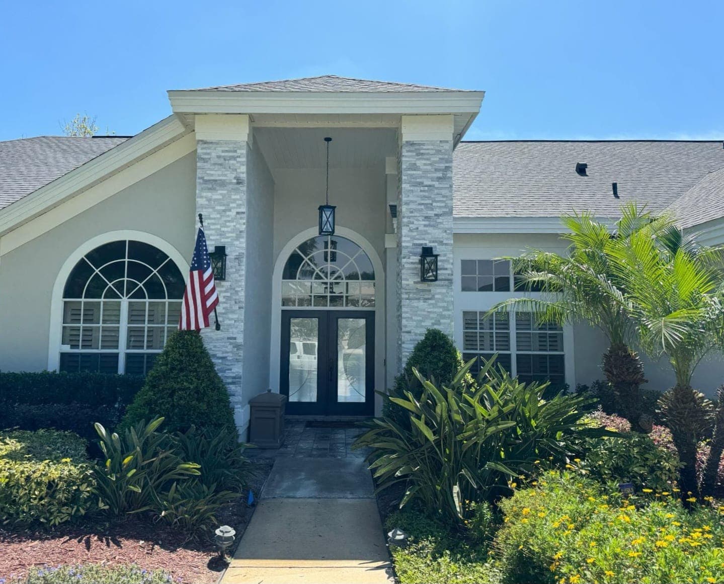 Modern home entrance featuring stone pillars, double doors, an American flag, and lush tropical landscaping.