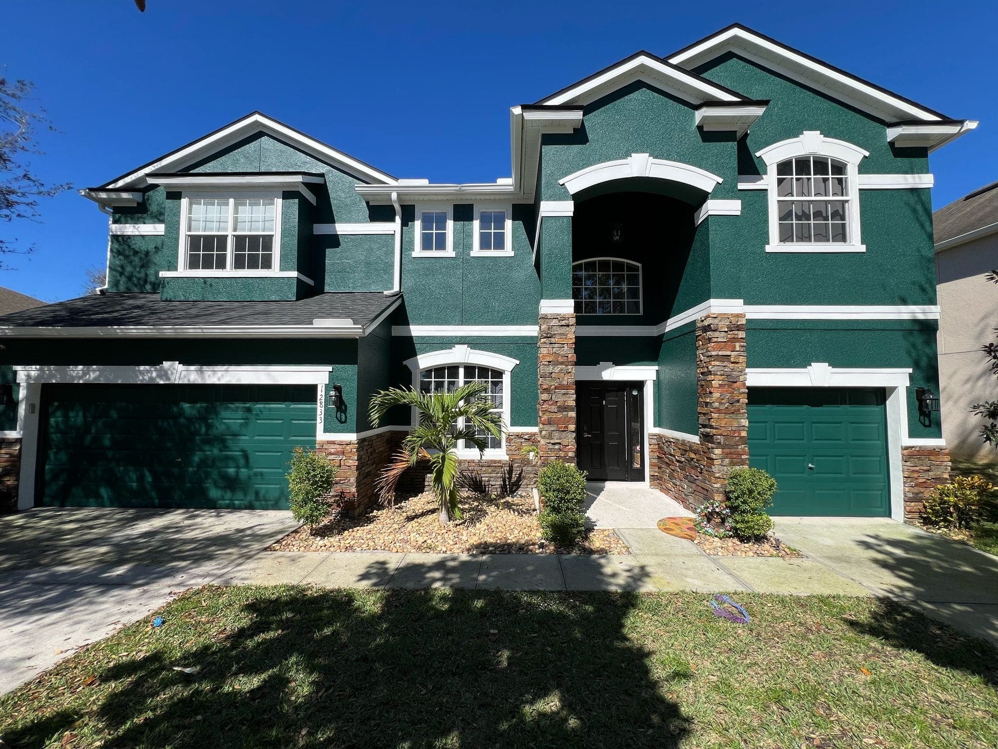 Two-story dark green house with white trim, stone accents, and two green garage doors.