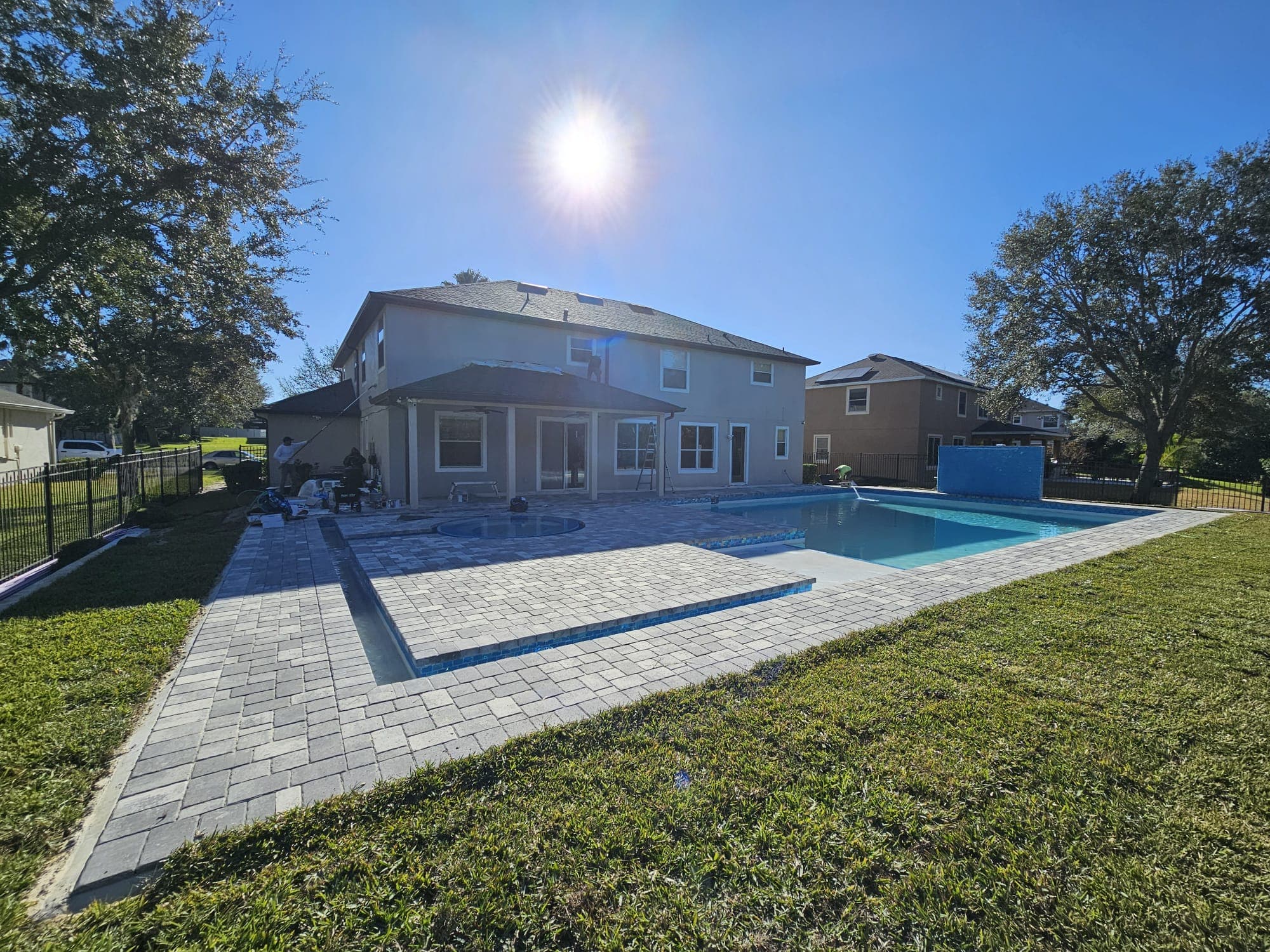 Stone paver patio and swimming pool behind a two-story house under a bright sun.