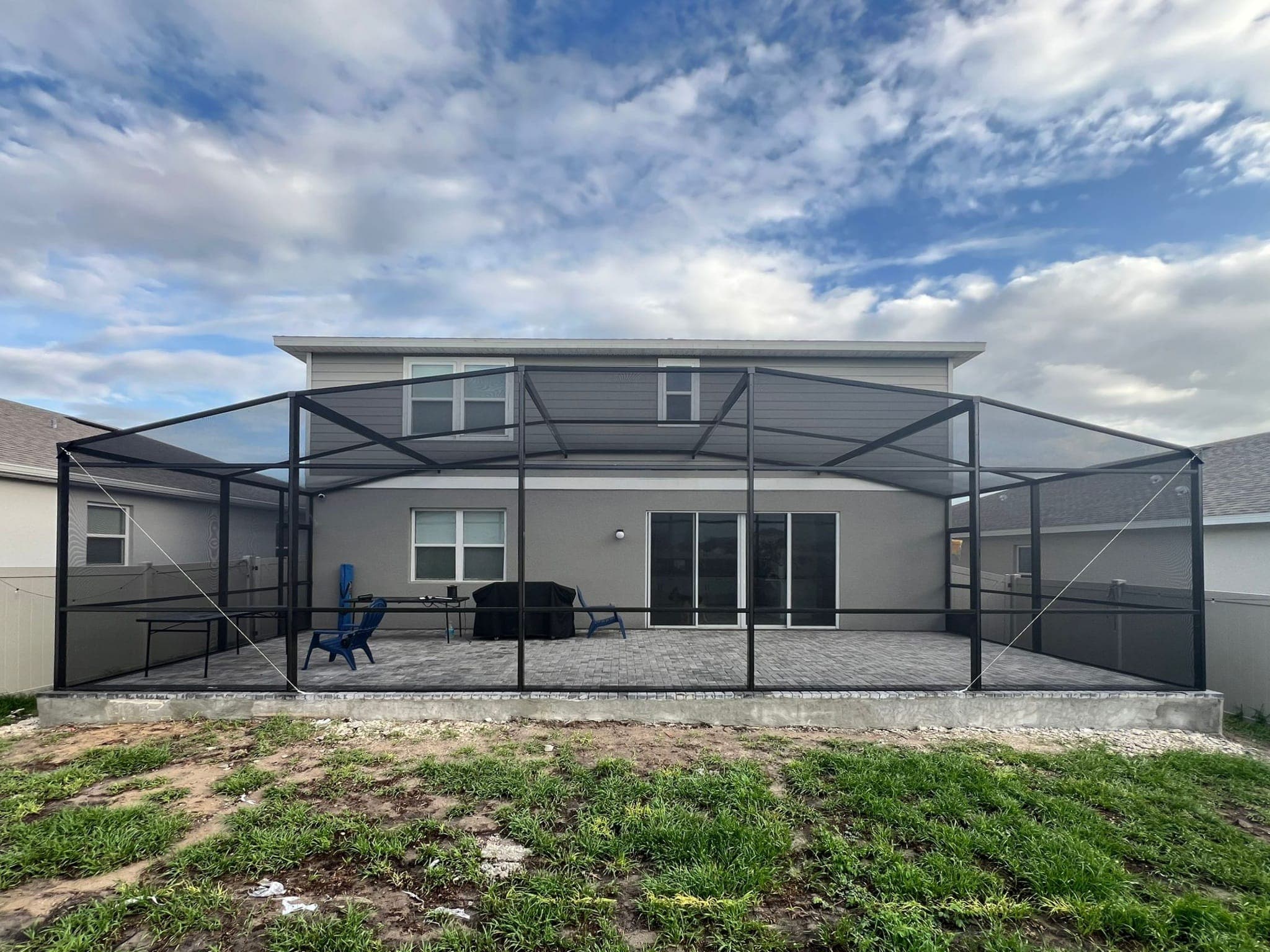 Large black screened-in patio enclosure attached to a two-story grey house with stone pavers.