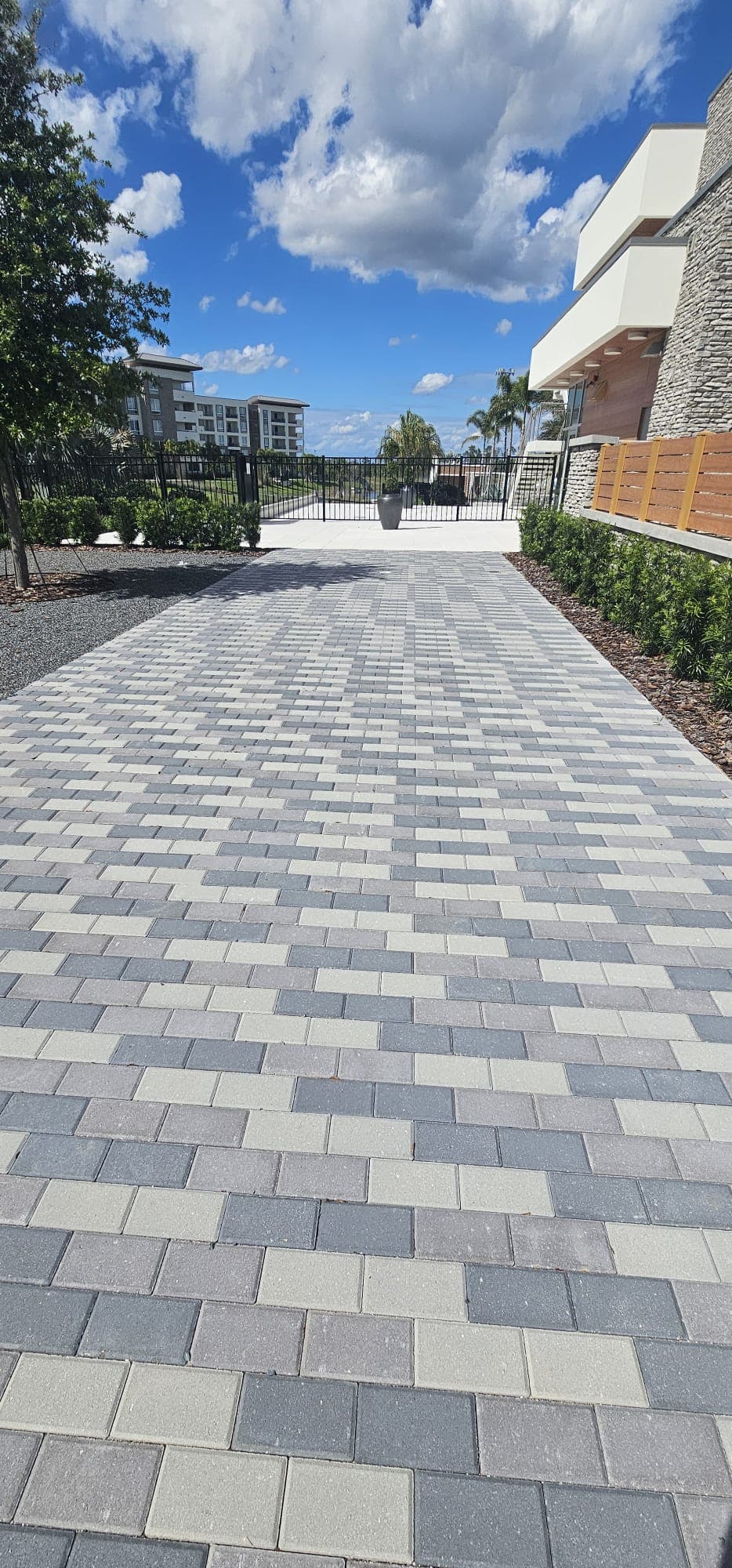 Gray and white paver walkway leading to a black fence under a bright cloudy sky.