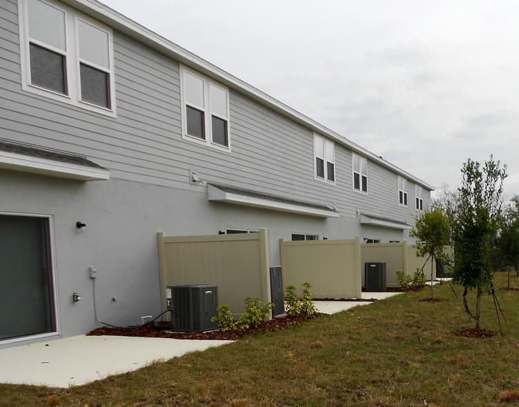 Rear of gray townhouses with concrete patios, air conditioning units, and tan privacy fences.