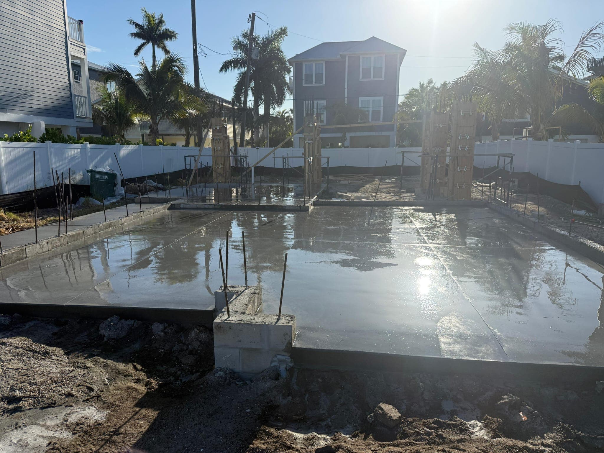 Wet concrete foundation reflecting bright sunlight at a residential construction site with palm trees.