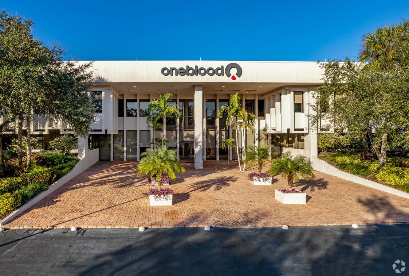 White OneBlood building featuring a brick courtyard and palm trees under a clear sky.